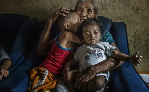 Remy Fernandez, 84-years-old, holds her grandchildren that she is raising, there are seven in all, because her son, Constantino de Juan, a methamphetamine user, was killed by masked men and the mother is in prison due to a drug arrest. The chair in which she sits has a hole in it after it passed through his body. Baby RJ was born in prison. Constantino, upon seeing the masked assassins, instructed CJ, 5-years-old and wearing the red tank top, to take care of his siblings because he knew he was about to be…