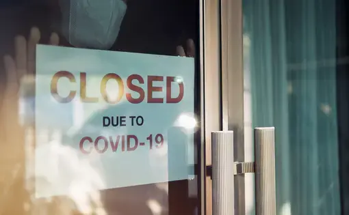 Business office or store shop is closed, bankrupt business due to the effect of novel Coronavirus (COVID-19) pandemic. Unidentified person wearing mask hanging closed sign in background on front door. Image by Chansom Pantip. Thailand, 2020.