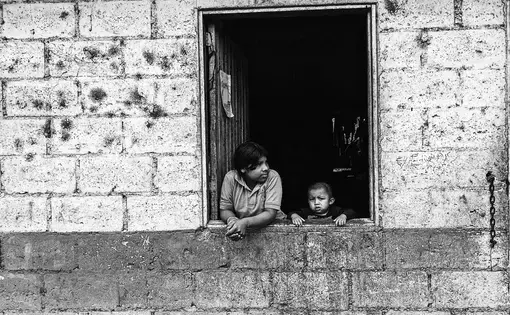 In a bache near the Rio Sereno border crossing between Panama and Costa Rica, a young boy and his older sister look out to the coffee fields from a lone window of their home. Image by Samira Tella. Costa Rica, 2018.