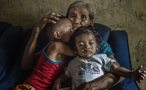 Remy Fernandez, 84 years old, holds two of the seven grandchildren she is raising. Her youngest son, Constantino de Juan, a methamphetamine user, was killed by masked men in December, 2016. Upon seeing his attackers, Juan instructed five-year-old CJ, shown here wearing a red tank top, to take care of his siblings. The children's mother is in prison due to a drug arrest. Baby RJ, in the "daddy's little helper" T-shirt, was born in prison. Image by James Whitlow Delano. Philippines, 2017.