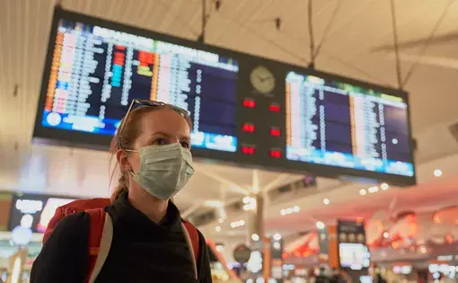 A woman with a backpack wearing an anti-virus mask on her face at the airport arrivals board. Image by Damian Pankowiec/Shutterstock. 2020.