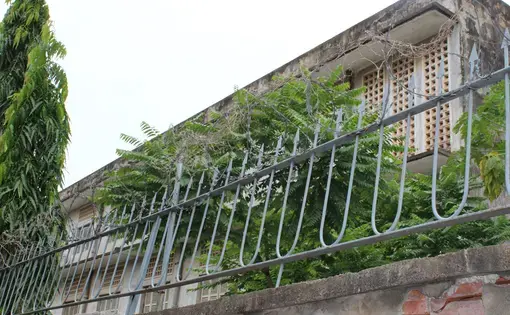 Photograph of S-21 former concentration camp, taken from the perimeter wall.  Barbed wires, spiked metal railings, and overgrown ferns partly shield the view of the building. Image by M.G. Zimeta. Cambodia, 2018.