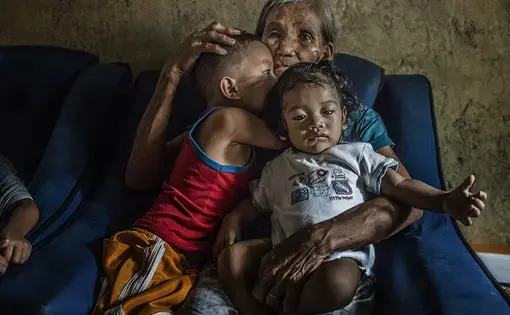 Remy Fernandez, 84 years old, holds two of the seven grandchildren she is raising. Her youngest son, Constantino de Juan, a methamphetamine user, was killed by masked men in December, 2016. Upon seeing his attackers, Juan instructed five-year-old CJ, shown here wearing a red tank top, to take care of his siblings. The children's mother is in prison due to a drug arrest. Baby RJ, in the "daddy's little helper" T-shirt, was born in prison. Image by James Whitlow Delano. Philippines, 2017.