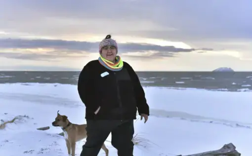 In this Jan. 14, 2019 photo, Clarice "Bun" Hardy stands on the beach with her dog, Marley, in the Native Village of Shaktoolik, Alaska. Hardy, a former 911 dispatcher for the Nome Police Department, says she moved back to her village after a sexual assault left her feeling unsafe in Nome. Image by Wong Maye-E. United States, 2019.