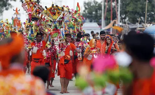 Hindu pilgrims, known as Kanwarias, walk near the banks of the Ganges River in Prayagraj, India, on July 25, 2019. Kanwarias are devotees performing a ritual pilgrimage in which they walk the roads of India, clad in saffron, and carrying ornately decorated canisters of sacred water from the Ganges River over their shoulders to take back to Hindu temples in their hometowns, during the Hindu lunar month of Shravana. Image by Rajesh Kumar. India, 2019.