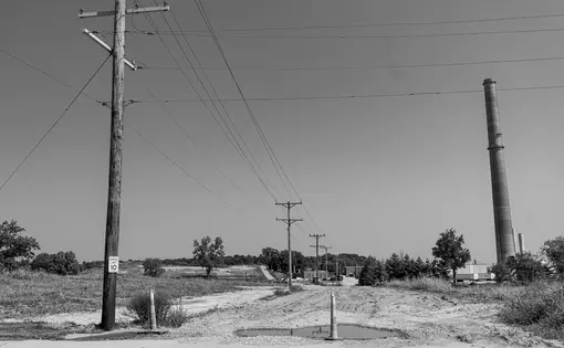 An old road covered in pot holes, sand, and dirt in front a clear sky. Telephone poles are lined down the street. There is a smokestack from the old lead smelter in the far right corner,