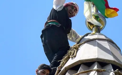 After climbing the temple cupola, a young Yezidi man hoists a flag and waves it to cheers from the courtyard below. Image by Kaitlyn Johnson. Georgia, 2019.