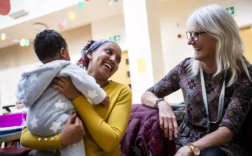 Eliad Abrha, 7 months, left,  is held by his mother Hanna Abrha, of Parkhead, a neighborhood of Glasgow, Scotland, center, as she speaks and jokes with Janet Tobin, health improvement lead with the National Health Service, right, at Cafe Stork, Tuesday, Oct. 21, 2019, at Parkhead Congregational Church in Glasgow. Cafe Stork is a free networking lunch for families in the disadvantaged East End who are expecting or new parents. Image by Michael M. Santiago. United Kingdom, 2019.