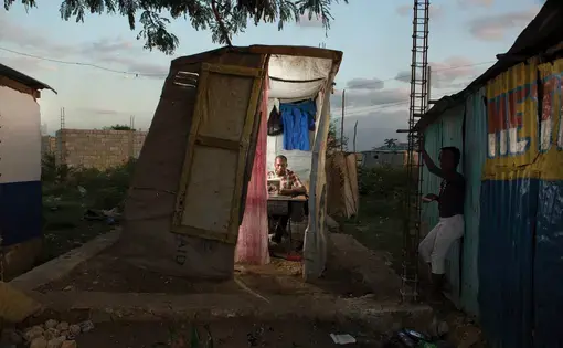 Reginald Adrasse repairs a uniform in his small self-built tailor shop at the entrance to Canaan 2. Image by Allison Shelley. Haiti, 2017.