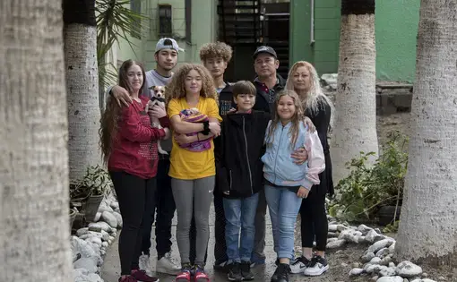 The Flores family pauses for a portrait outside Ramon’s apartment complex in Tijuana, Mexico. Family members are, clockwise from left, Leslie, 23, Lennes, 18, Raymond, 15, Ramon, 46, Enedis, 54, Rayma, 10, Edward, 12, and Kennedy, 16. The dogs are Canelo, left, and Bambi, wrapped in the blanket. Image by Amanda Cowan. Mexico, 2019.
