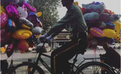 A member of the Chinese military rides his bike past two men selling balloons on Sunday, Sept. 24, 2017, on the same block as the residence of U.S. Ambassador to China Terry Branstad in Beijing, China. Image by Kelsey Kremer. China, 2017.