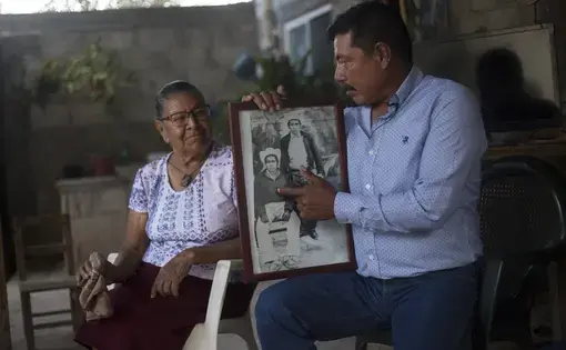 Winny Contreras sits with his mother, Cecilia Galvan, in his childhood home. He points to an old photo of his father who also worked on farms in the United States. Image by Ingrid Holmquist. Mexico, 2018.