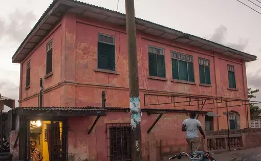 Tailor Pierre Kwetey inside his workshop, as a man walks by the house that allegedly contained a fake US Embassy in Accra. Image by Yepoka Yeebo. Ghana, 2017.