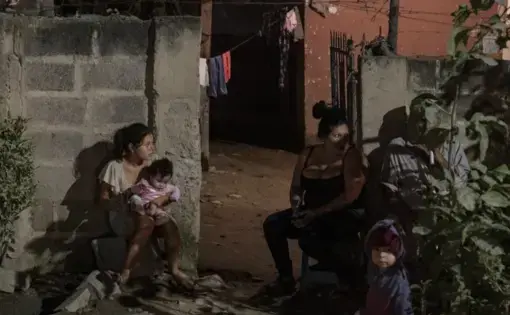 A family sits outside their home as forensic workers investigate a body at a crime scene in the Rivera Hernandez neighborhood of San Pedro Sula, Honduras on Nov. 30, 2019. MS-13. Mara 18. Los Vatos Locos _ The Crazy Guys. The gangs' shifting lines of control dodge and weave through Rivera Hernandez, a place where even the police are afraid. Image by Moises Castillo/ AP Photo. Honduras, 2019.