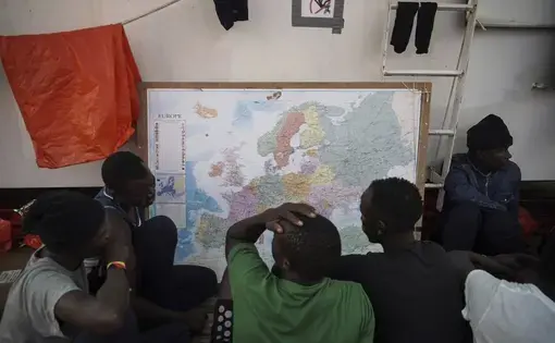 In this Sept. 23, 2019 photo, rescued migrants look at a map of Europe aboard the Ocean Viking humanitarian ship as it sails in the Mediterranean Sea. The misery of migrants in Libya has spawned a thriving and highly lucrative business, in part funded by the EU and enabled by the United Nations, an Associated Press investigation has found. Image by Renata Brito/AP Photo. Libya, 2019.