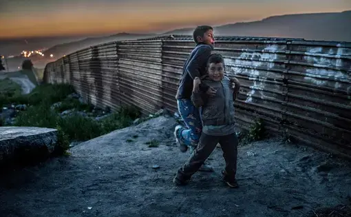 Two boys from the Arias family roughhouse in the dust beside their home abutting the original U.S.-built border wall in Colonia Libertad, Tijuana, Mexico. Behind them, a string of lights set up by U.S. Border Patrol stretches to the sea. The lights were set up by U.S. Border Patrol to illuminate the wall to make it more difficult migrants to cross at night. Image by James Whitlow Delano. Mexico, 2017.