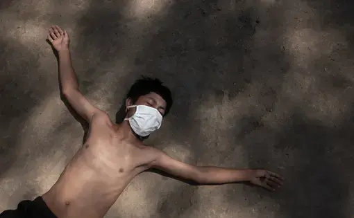 Image by Florence Goupil. Peru, 2020. A young boy lies down on the dirt ground with arms outstretched. He is wearing a white surgical mask, black shorts, and no shirt.