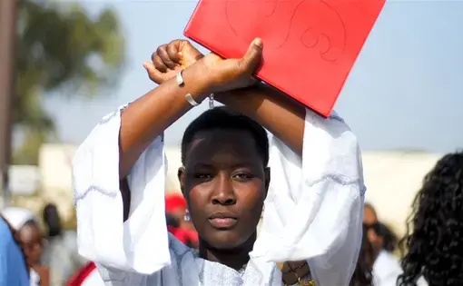 Image by Ricci Shryock. Senegal, 2012. Student dressed in white makes an X with arms above head while holding a red folder