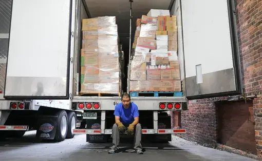 A truck driver leans on the edge of his 18-wheeler, which is full of boxes.