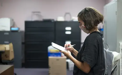 Carly Berlin, wearing a mask, stands and writes in a notebook.