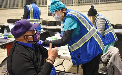 A man in a purple mask and baseball cap is sitting and giving a thumbs up as a woman in a mask and beanie administers a shot to his arm.