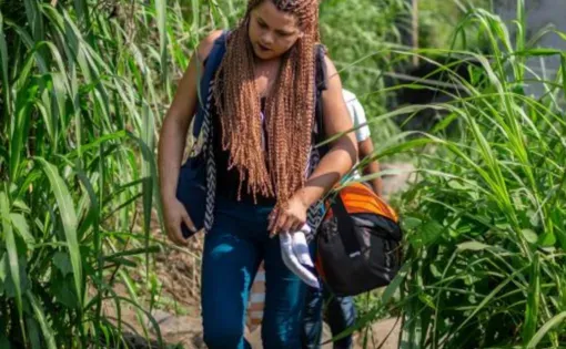 A young woman with long braids walks down a stone path with foliage on either side of her. She's wearing jeans and sandals and carries with her a duffel bag and a purse.