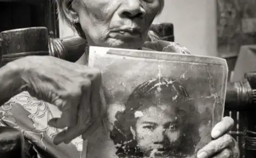 A black-and-white image of an elderly woman holding a photo of herself as a young girl