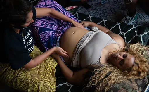 Overhead shot of a pregnant woman lying on a bed with her eyes closed, her head at the bottom right of the frame, and her stomach at the top left of the frame. Her tank top is rolled up, and someone to her left is placing their hands on either side of her stomach.