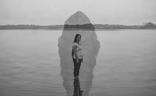 Black and white portrait of a woman standing in a lake, with the water coming up to her ankles. She has long dark hair and wears a white shirt and dark pants. Superimposed on top of her is a semi-translucent image of a leaf.