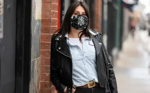 Portrait of Allison June Palmer on the sidewalk leaning against a brick wall. She's wearing a leather jacket and a black mask with an image of a skeleton on it.
