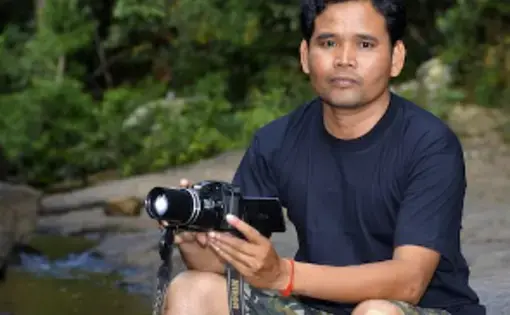 Portrait of Leng Ouch holding a camera and sitting on a rock next to a lake with trees behind him.