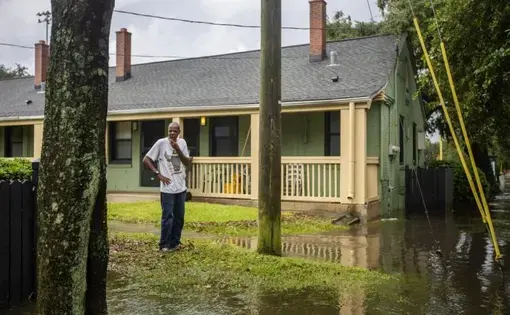 A man stands in the grass in front of a one-story residential building. The ground is flooded around him, and he surveys the scene with a hand on his chin.