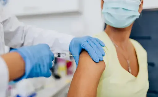 A medical professional wearing gloves inserts a needle into a masked woman's arm