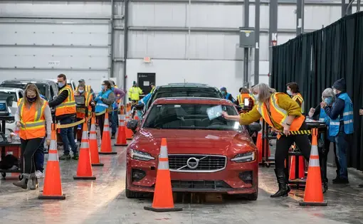 Workers in masks and reflective vests distribute vaccines as people wait in cars.