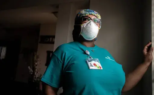 A woman stands with one hand against a wall. Her hair is covered with a scrub cap and she's wearing a mask and a teal shirt with a badge pinned to it.