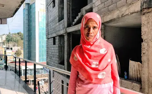 A portrait of an Ethiopian woman standing on a balcony. She is wearing a pink, long-sleeved shirt and a pink scarf with white polka dots covers her hair.