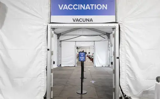 View of the entrance of a white tent. Above the entrance is a sign that says "vaccination" and "vacuna." In the entryway is a post with a sign on top directing patients to go to the right and staff to go to the left. There are white arrows on the ground to direct traffic.