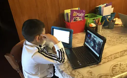 A young boy sits at a kitchen table with a tablet and laptop in front of him. On the laptop, he has a Zoom meeting open with four other attendees. On the table, there are three cups holding writing utensils and red, green, and blue containers holding notebooks and papers.