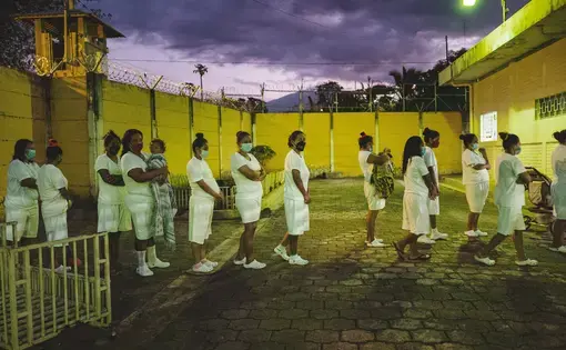 LOBA-ANAMARIA-AREVALOGOSEN-DIASETERNOS-0018.jpg Waiting to go inside, a line of women stand in a yard enclosed by a wall that is topped with barbed wire. The women wear the same all-white uniform of a t-shirt, shorts, and crocs, and they all wear masks. Two women are holding small children. On the right of the frame is a stroller.
