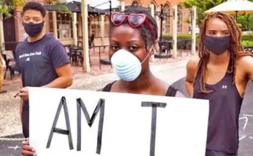 Hayes, wearing a medical face mask, holds up a sign that reads "Am I next?" Two other young people stand in the background.