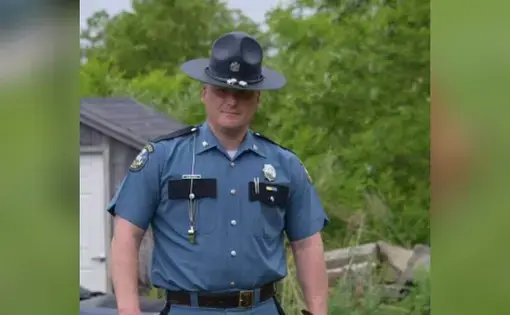 A man in a state trooper's uniform and hat stands outside. Behind him are trees.