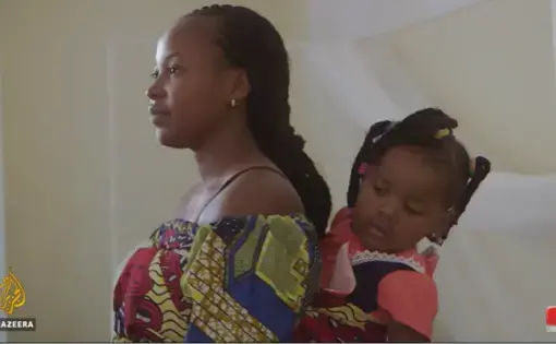 A Congolese mother carries her young daughter on her back in a bright, patterned sling. She is standing in a doctor's office in front of a small white bed.