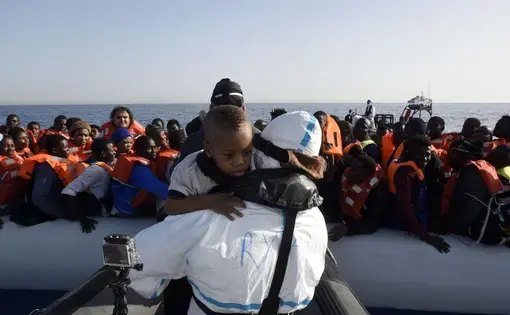Dozens of Africans are crammed in a raft, all wearing life vests. Beyond them is the sea. In the foreground in front of the raft, someone wearing a white, waterproof uniform is carrying a little boy who was presumably on the raft. The boy looks over the person's shoulder, toward the camera.