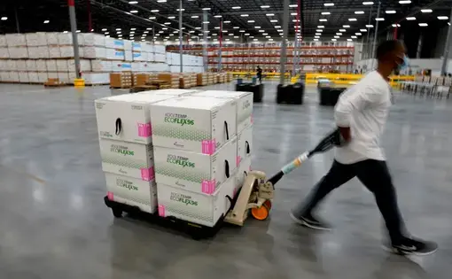 A man in a mask and white shirt pulls a pallet carrying 12 boxes of the Moderna vaccine. He is in a warehouse, and behind him are shelves filled with countless other boxes.