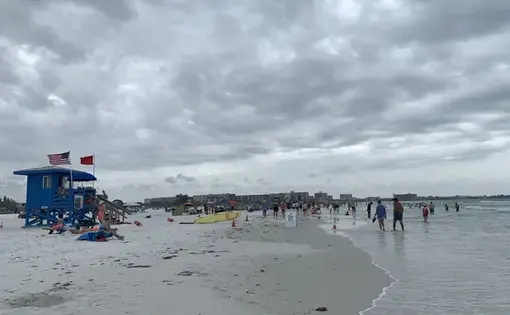 Image of beach with a blue lifeguard tower on the left of the frame and the ocean on the right. Dozens of beachgoers sit on the sand or walk through the tide. A few carry surfboards.