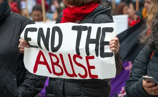 Female protester with a banner that reads "end the abuse." She has short brown hair, and her face is turned away and mostly out of frame. She's wearing a black puffer coat and a red scarf.