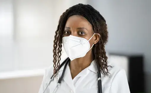 A woman wearing a white medical mask and a white uniform with a stethoscope around her neck.