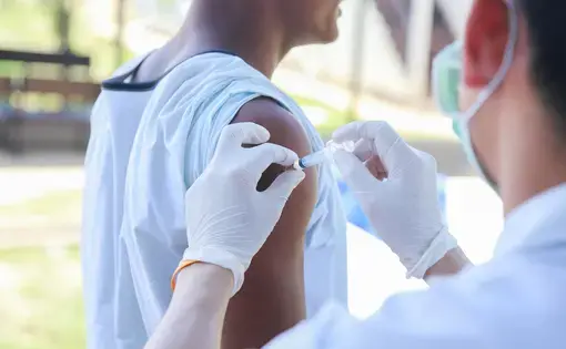 A man with the sleeve of his blue shirt rolled up is injected by someone mostly out of frame who is wearing gloves and a mask.