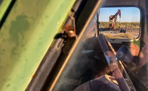 The camera is looking into the driver's side window of a vehicle. Through the passenger's side window, we can see an oil pump jack fracking crude extraction machine.