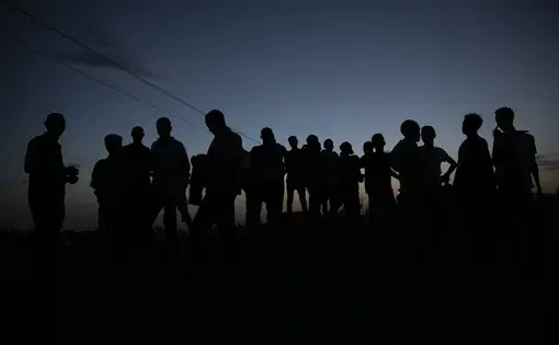 About 20 men stand outside at nighttime. We can only see their silhouettes against the night sky.
